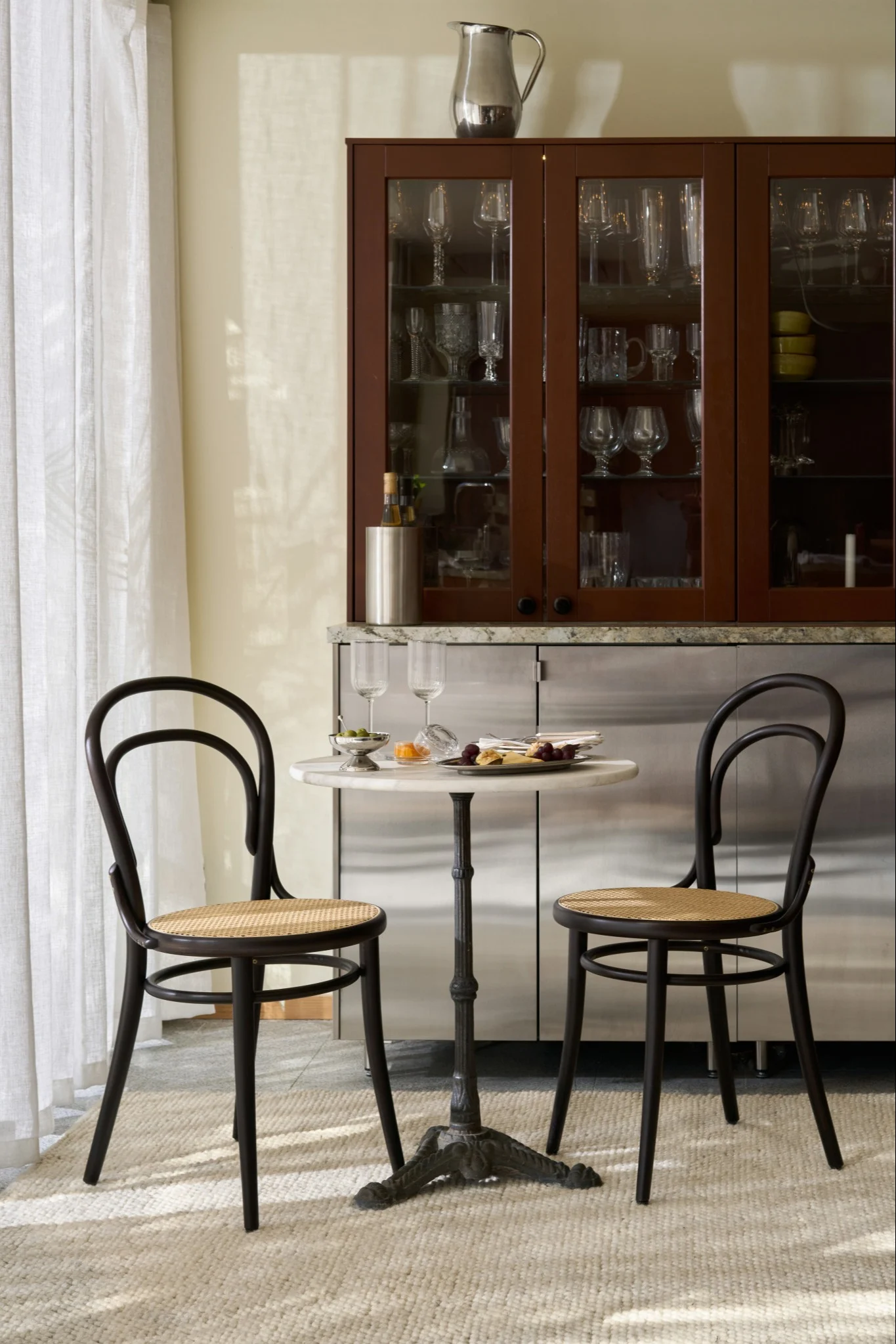 Two dark bentwood chairs and a small marble table with snacks in front of a bar cabinet with glassware.