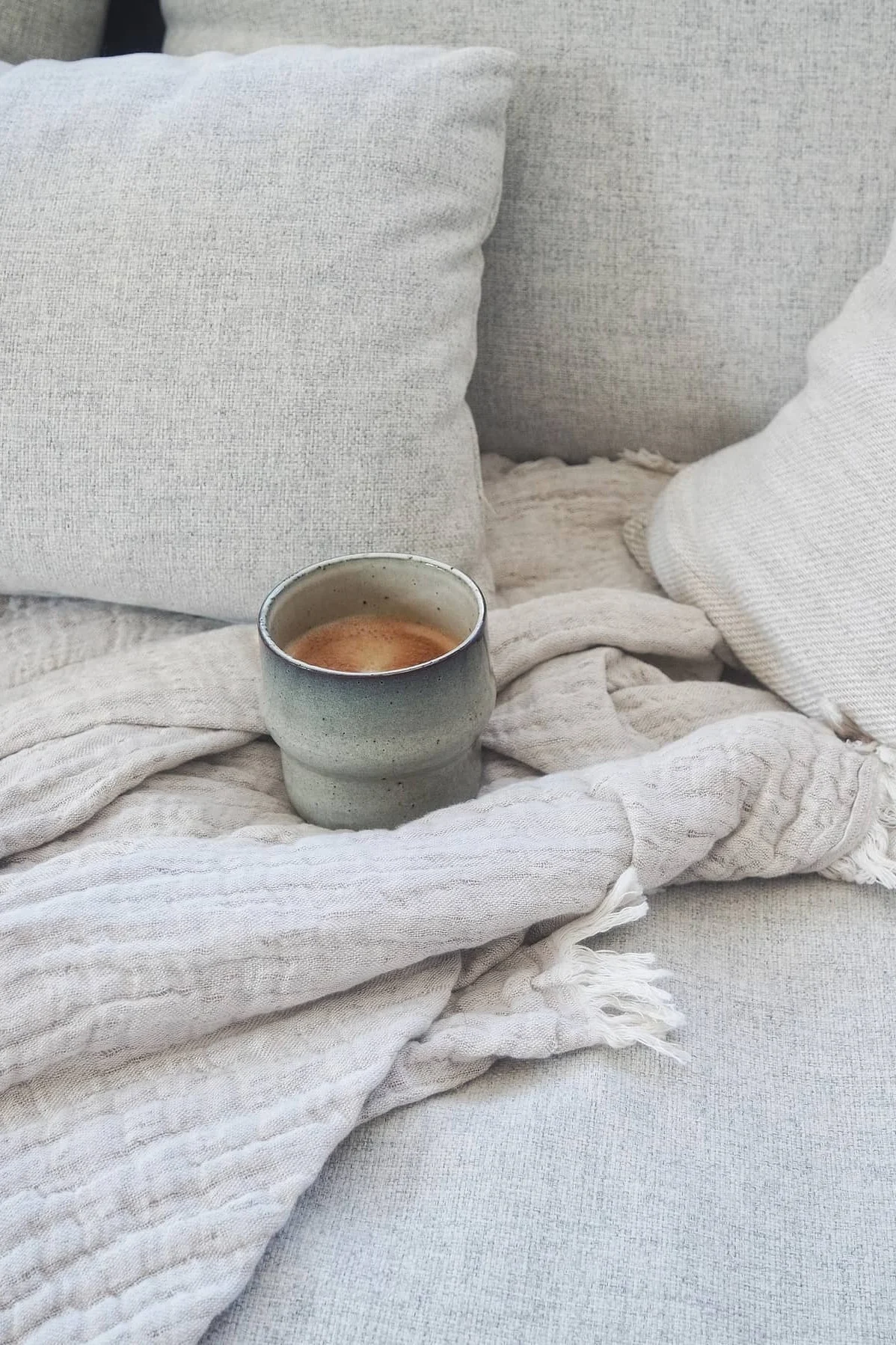 A ceramic mug with coffee rests on a light textured blanket and sofa amidst pillows.