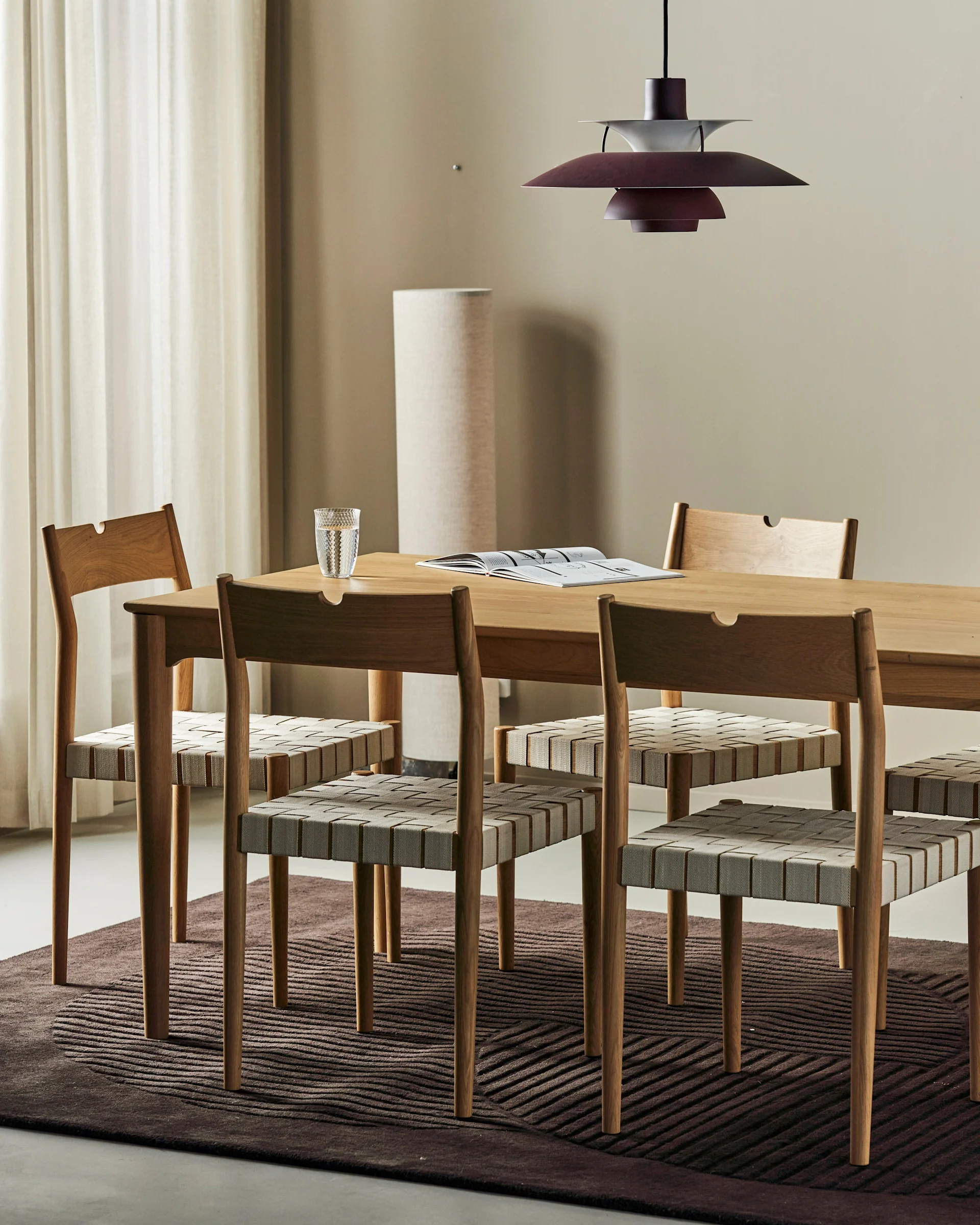 A modern dining room with a wooden table, six chairs with woven seats, a textured brown rug, and a multi-tiered red hanging lamp.