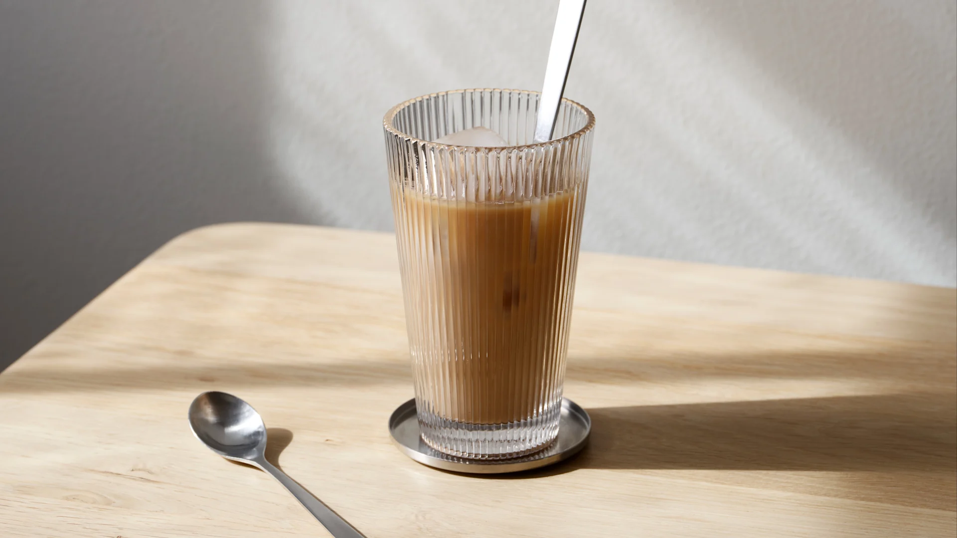 Iced coffee in a ribbed glass with a spoon on a wooden table, with sunlight and shadows.