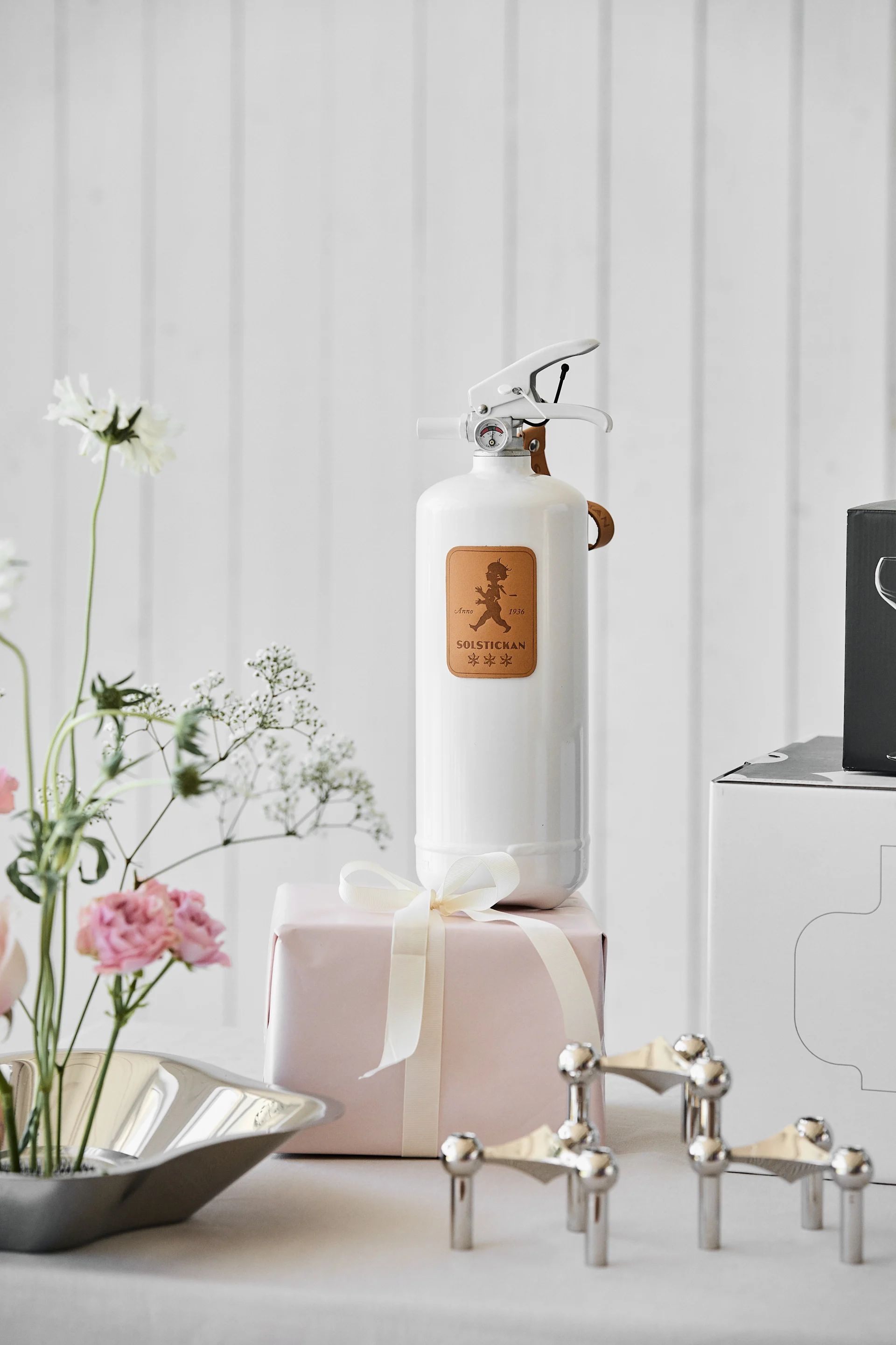 White fire extinguisher with a brown label featuring a figure and text, standing on a pink gift box with a ribbon, surrounded by flowers.