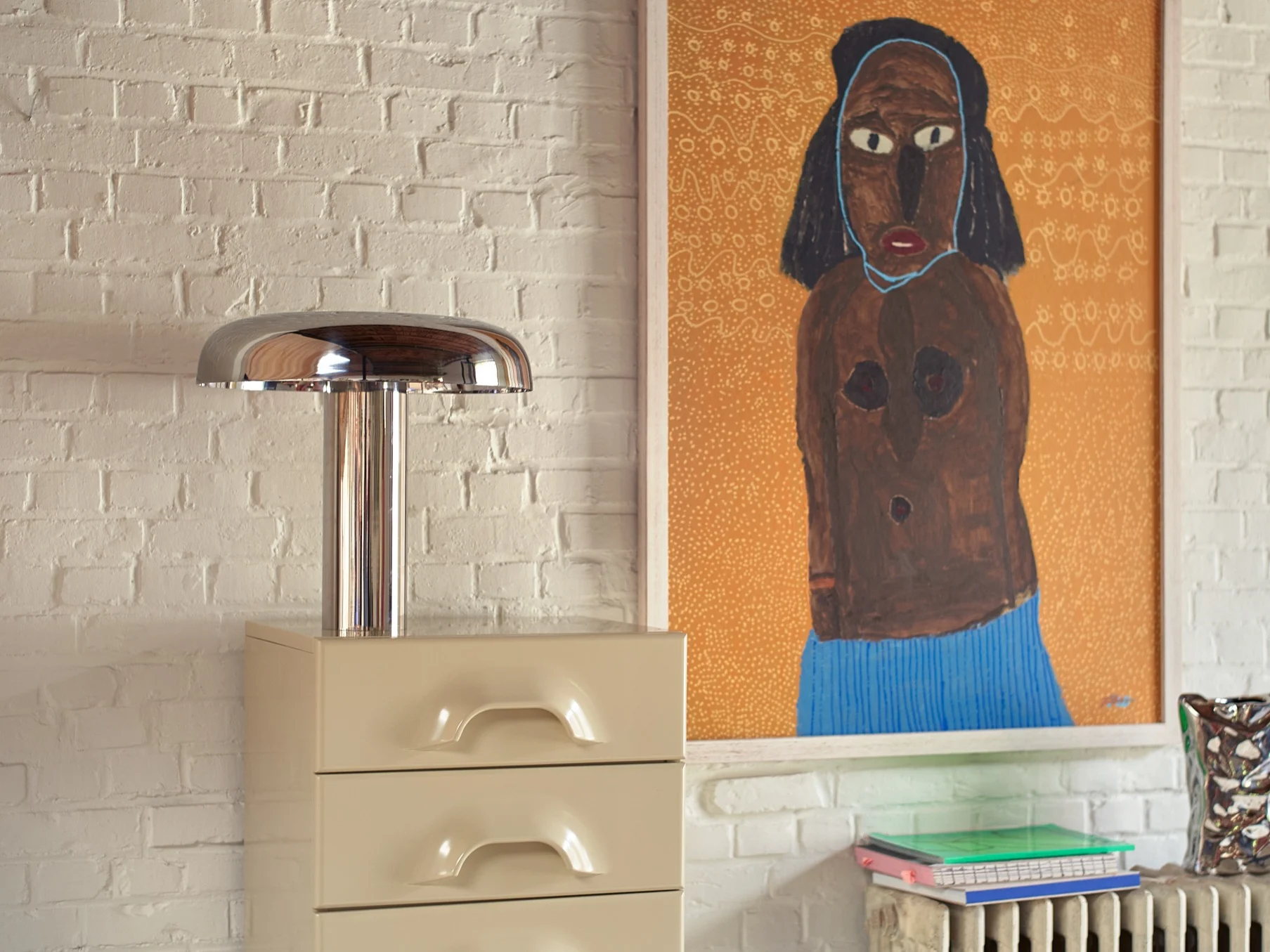 A chrome mushroom lamp on a beige dresser, next to a framed painting of a figure, with notebooks and a vase.