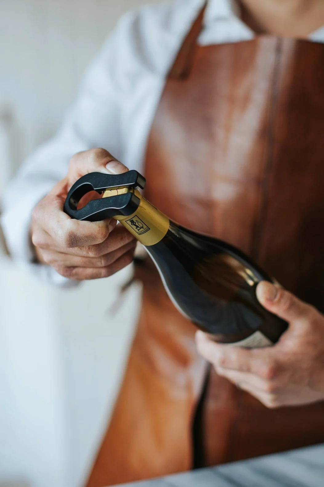 A person in a brown apron uses a black bottle opener to remove the cork from a sparkling wine bottle.