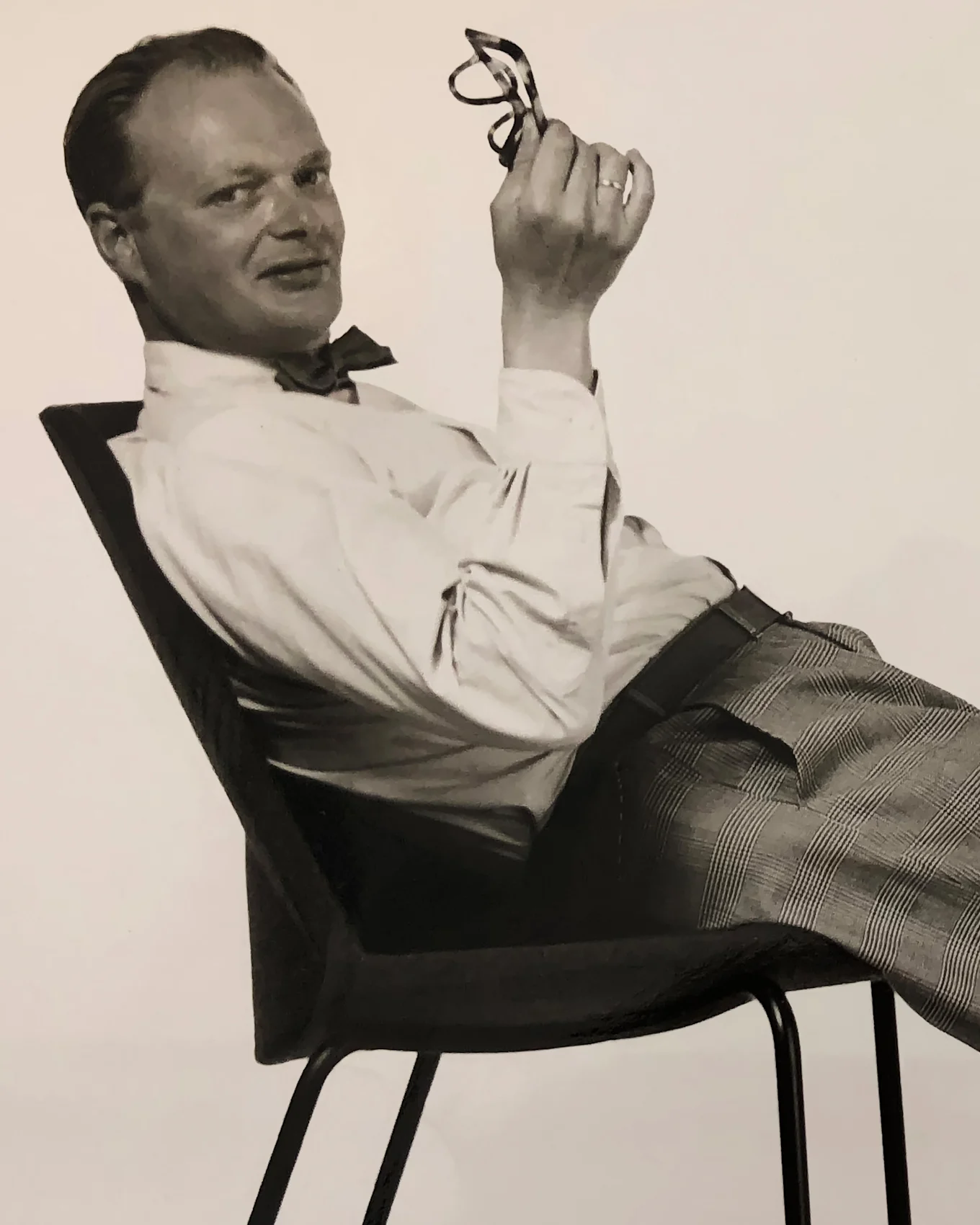 Man in bow tie and plaid trousers leaning back in a chair, holding eyeglasses, smiling at the camera.
