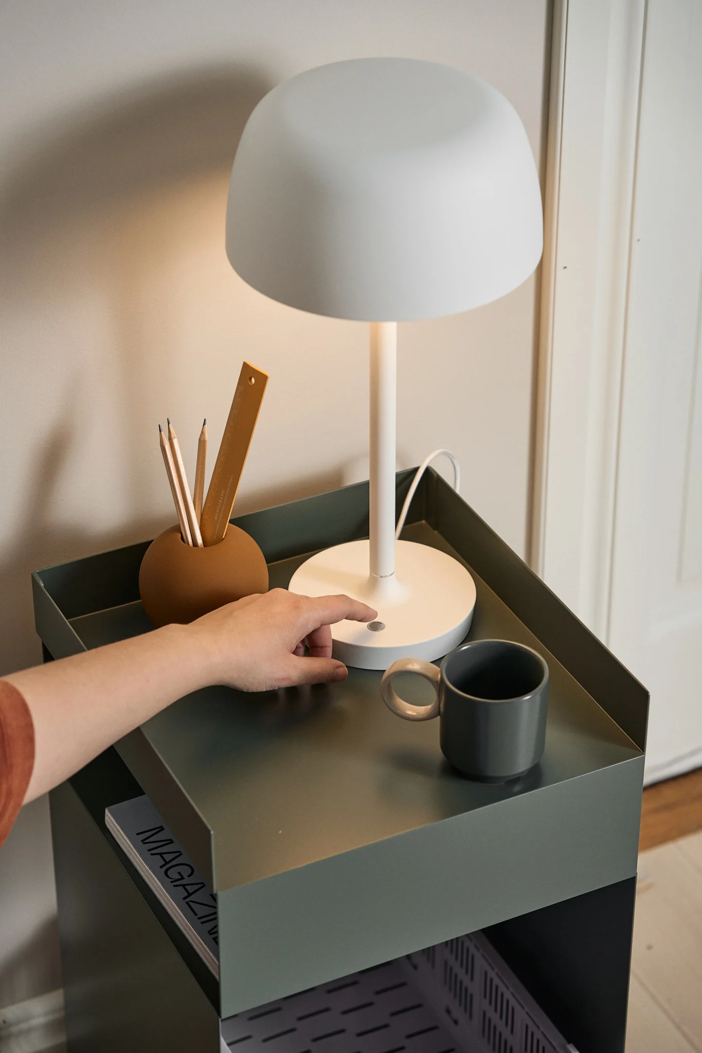 A hand switches on a modern white table lamp on a dark green metal side table with a pen holder and mug.