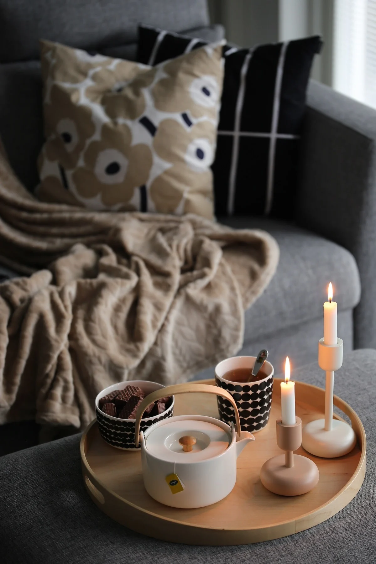 A cozy scene with a wooden tray on a grey sofa, holding a white teapot, a black and white patterned teacup, chocolates, and two lit candles.