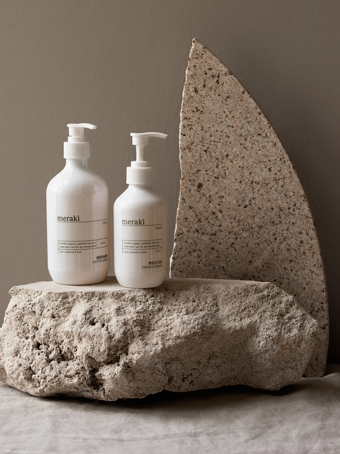 Two white Meraki pump bottles, a bodywash and a multi oil, stand on rough light-colored stones against a beige background.
