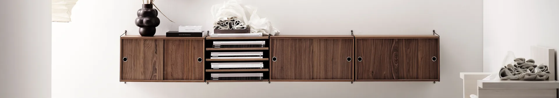 Wall-mounted wooden cabinet with sliding doors, open shelves, a sculptural dark vase, and stacked books.