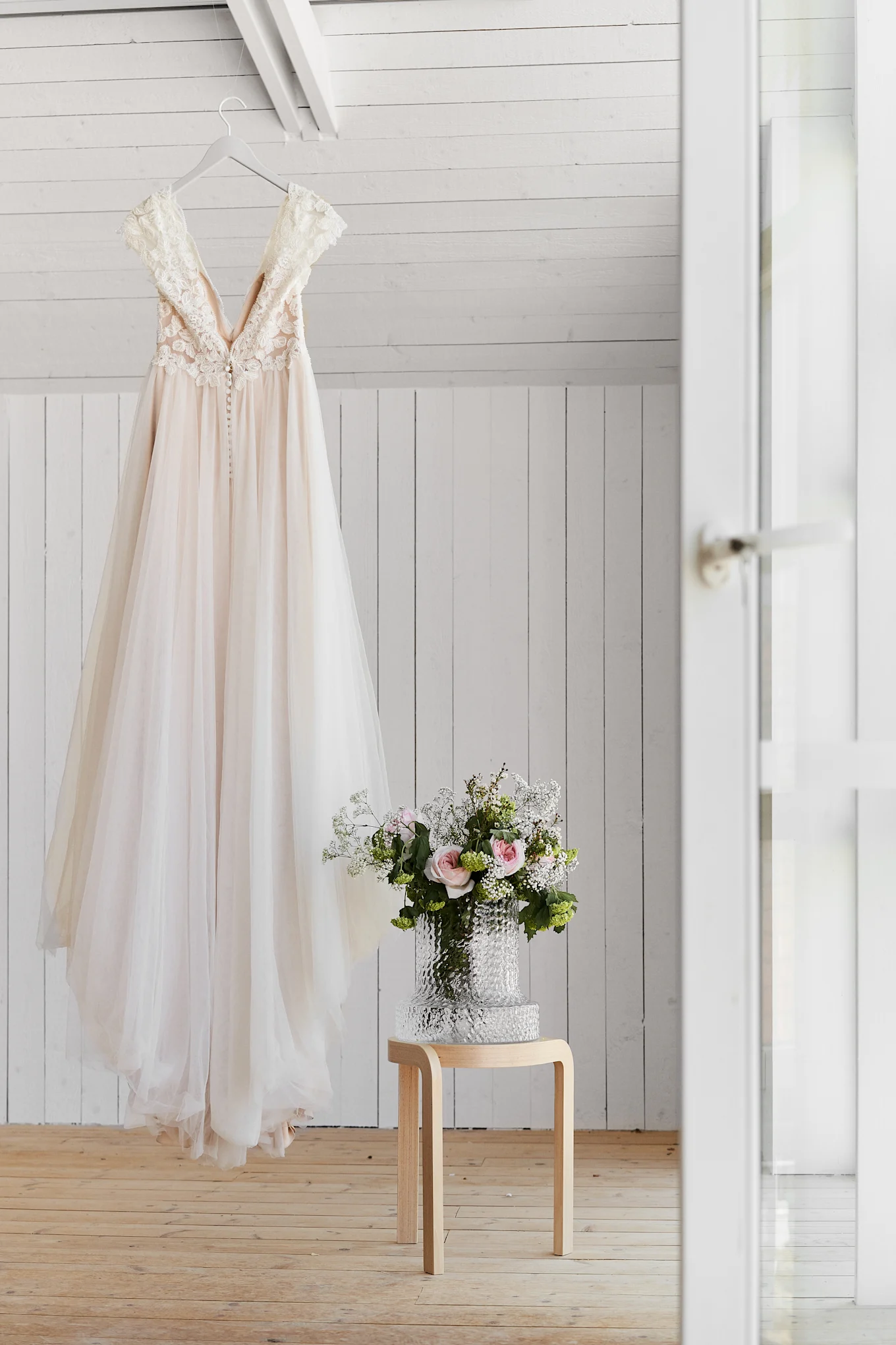 A light peach wedding dress with lace detail and a buttoned back hangs on a hanger. A bouquet of pink roses and white flowers is on a wooden stool.