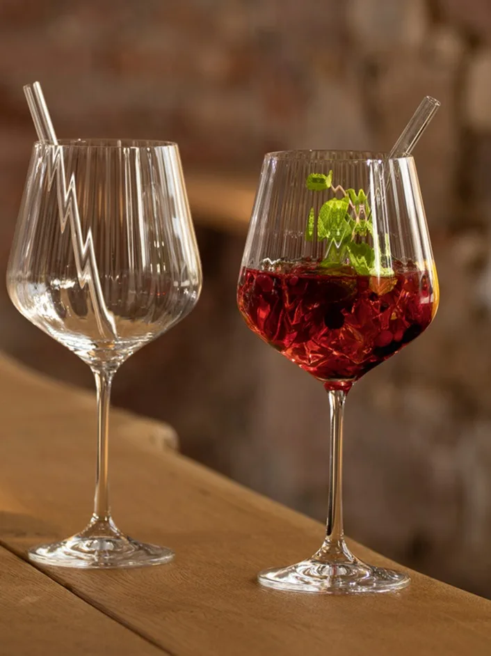 Two tall, ribbed stemmed glasses with straws on a wooden surface, one empty, the other with a red drink and mint.