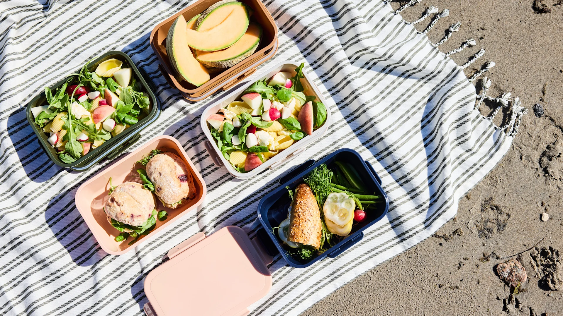 Picnic spread on a striped blanket on sand, with multiple lunch boxes containing fruit, pasta salad, and sandwiches.