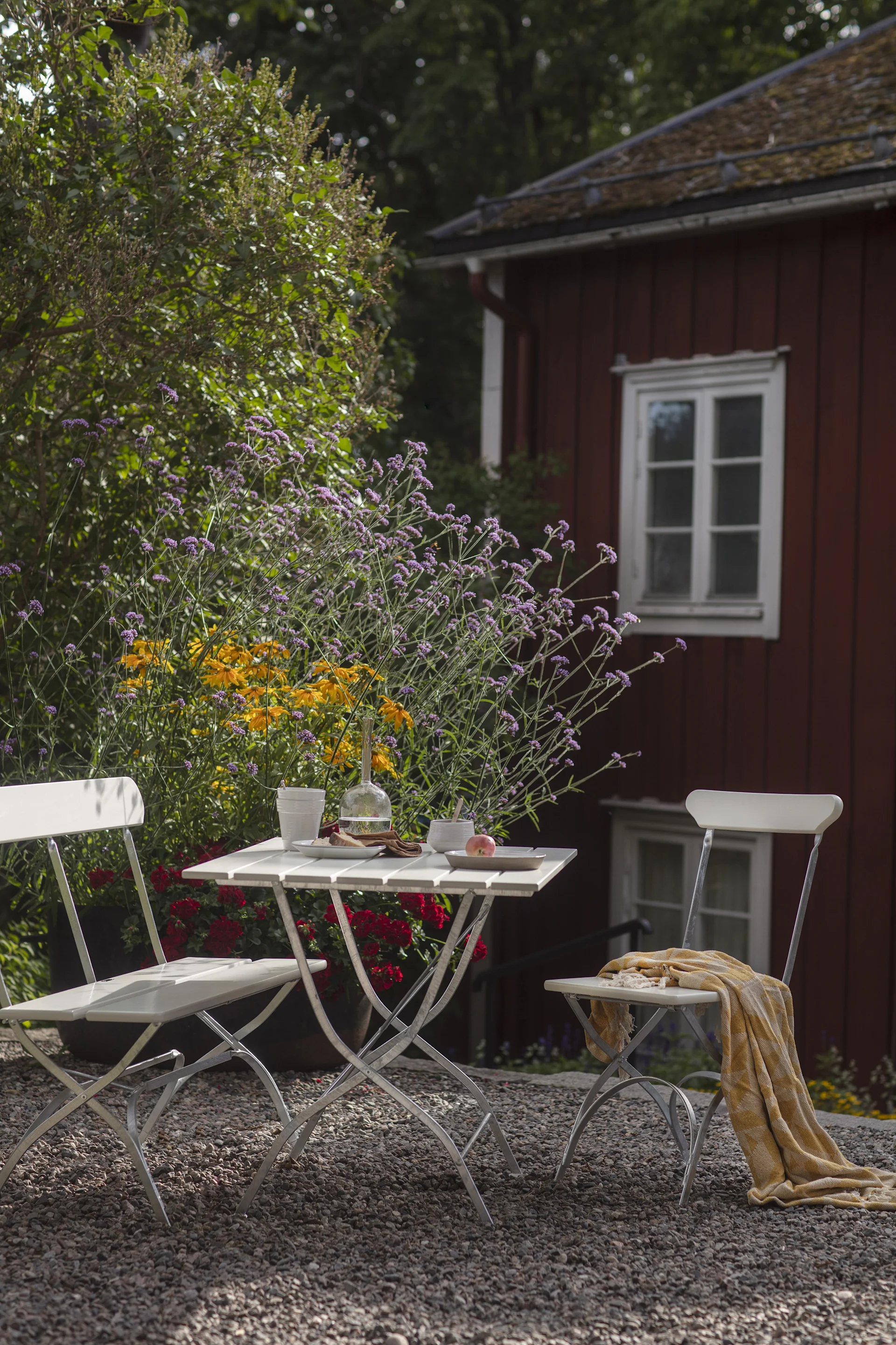 Outdoor bistro set with white table and chairs on gravel, laden with drinks and snacks, surrounded by vibrant flowers and a red house.