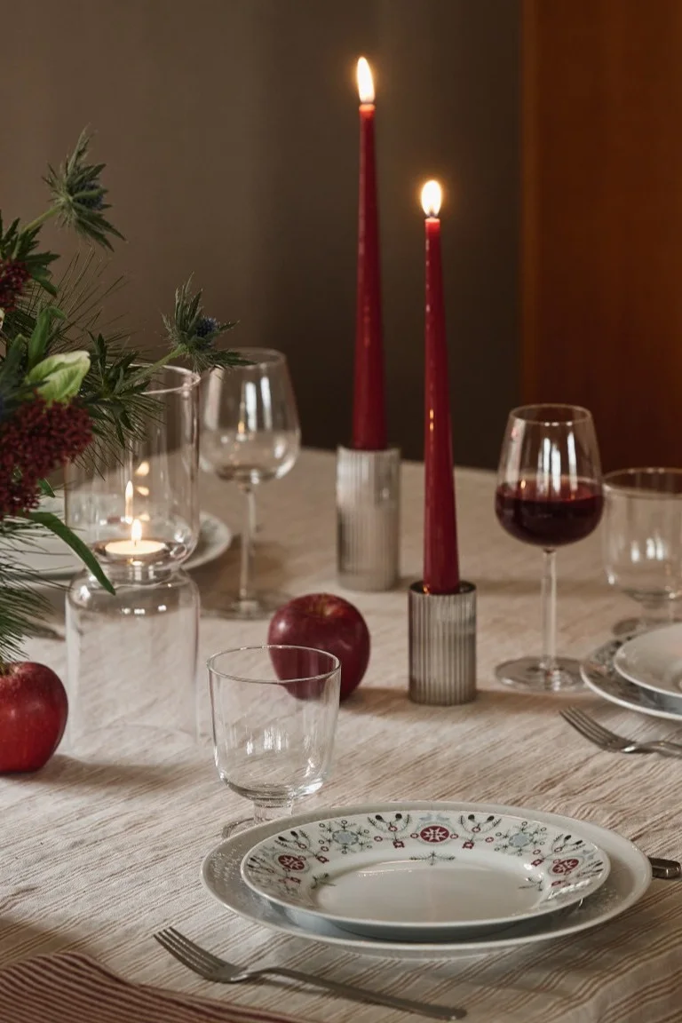 A dining table set with striped tablecloth, lit red tapered candles, wine glasses, patterned plates, and decorative red apples.