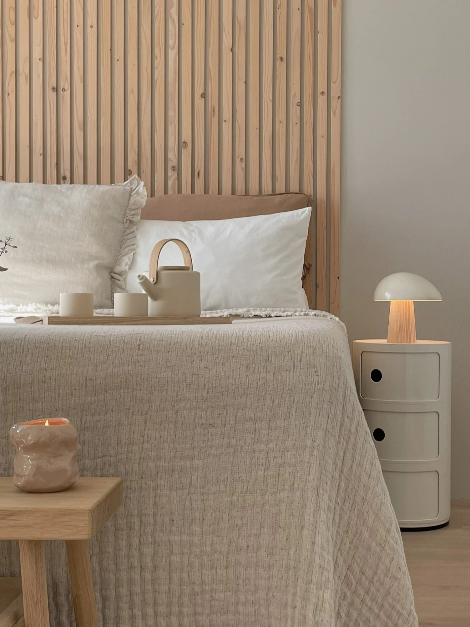 A neutral-toned bedroom with a wooden slatted headboard, a bed with textured bedding, a tea set, a mushroom lamp, and a candle on a stool.