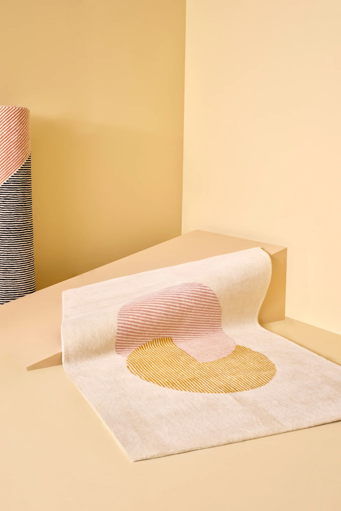 A light beige rug with abstract pink and yellow striped shapes, displayed on a beige geometric background next to a rolled striped fabric.