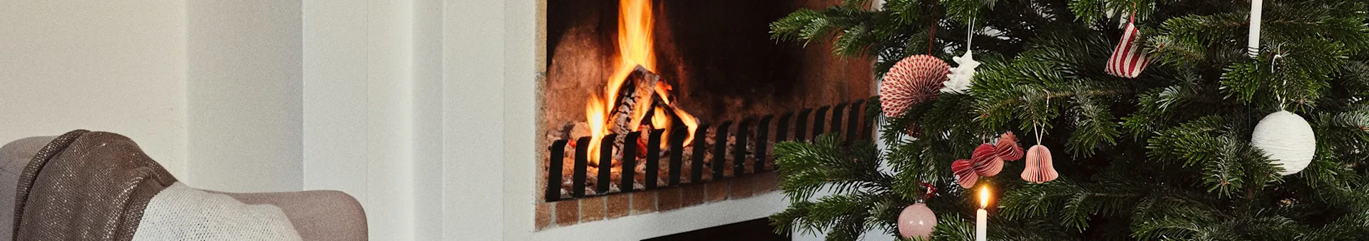 A festive Christmas tree with ornaments and a lit candle next to a warm fireplace with burning logs.