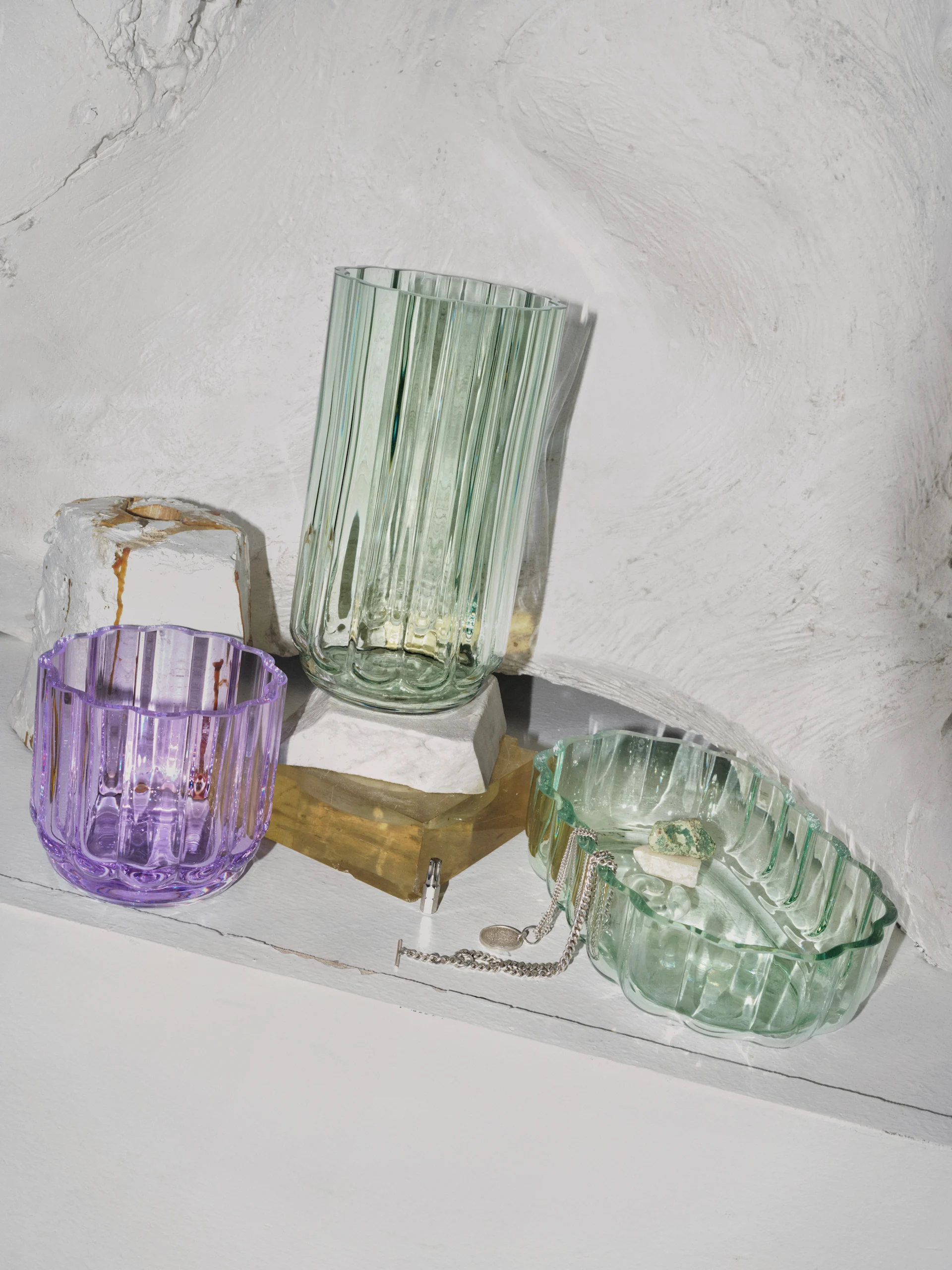 Three ribbed glass vessels: a purple cup, a tall green vase, and a green dish with a necklace and stones, on a white shelf.