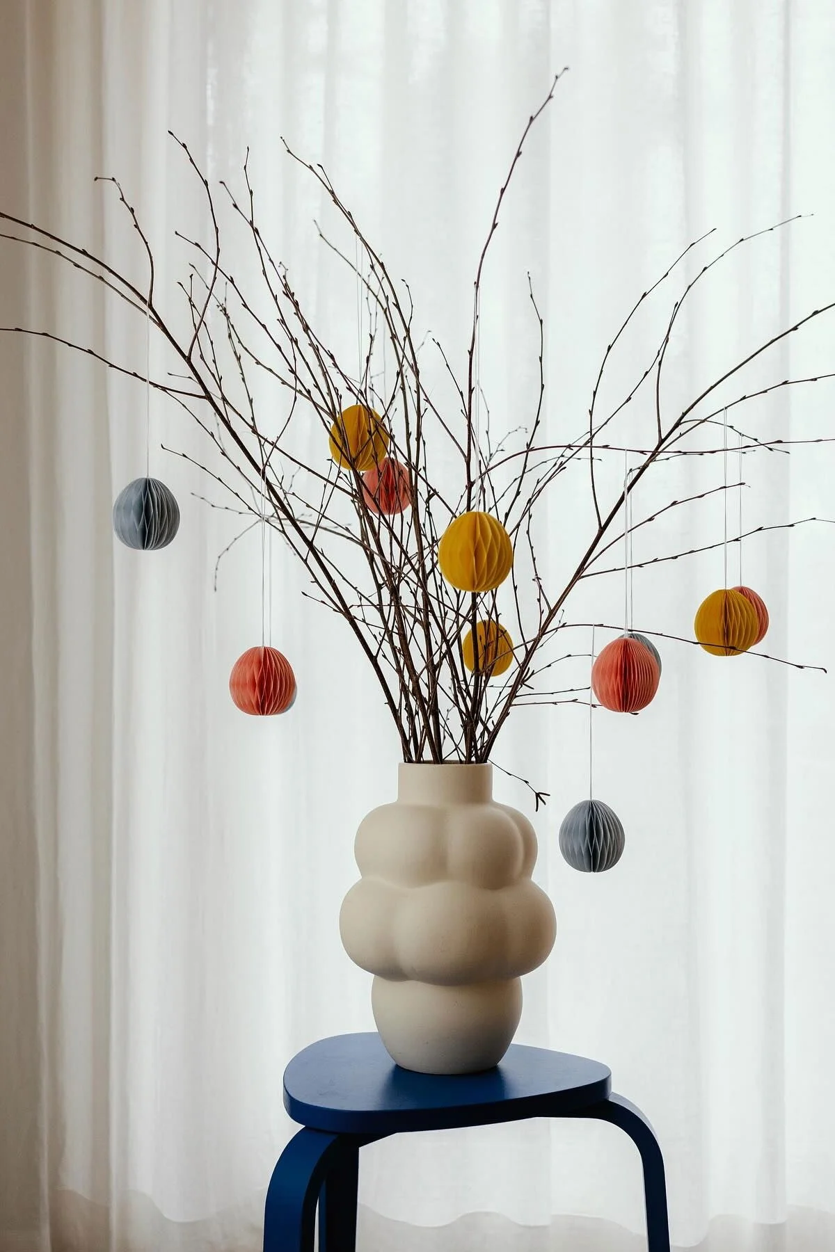 Cream vase with bare branches adorned with colorful paper egg ornaments on a blue stool.