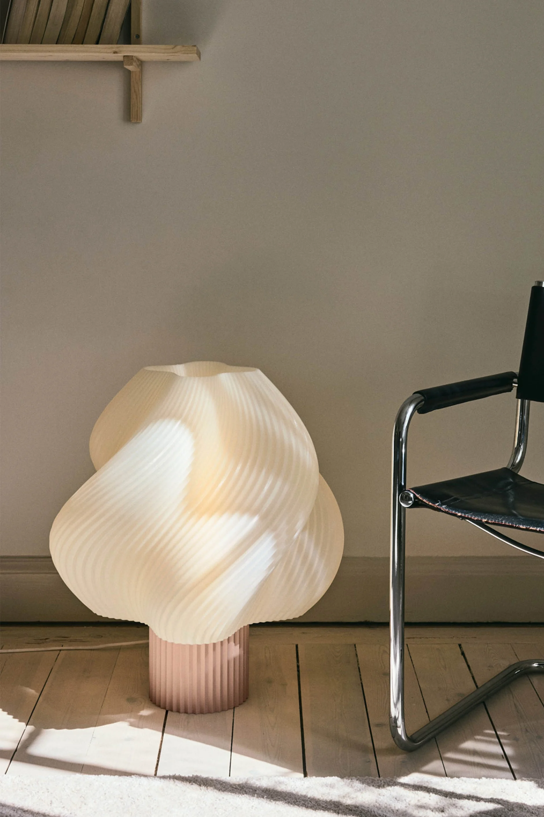 A sculptural, pleated cream floor lamp with a pink base stands on a wooden floor next to a black leather and chrome chair.