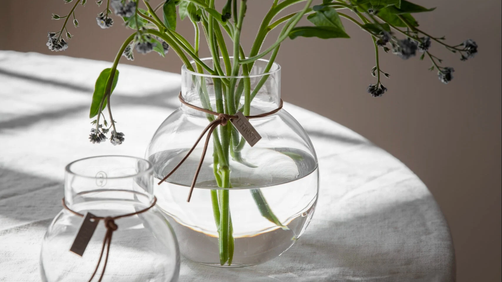 Two clear glass vases on a white tablecloth. The larger vase holds flowers and has an 'ERNST' tag.