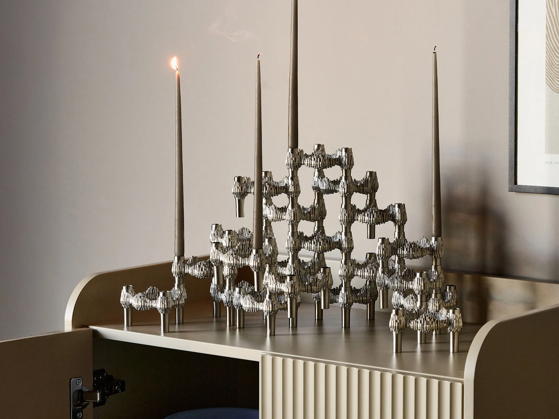 A textured, modular silver candelabra holds four grey candles: one lit, one smoking, and two unlit, on a light cabinet.