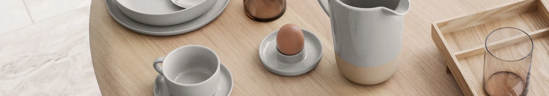 Light grey ceramic tableware, egg cup with egg, pitcher, and glass on a wooden table with a tray.