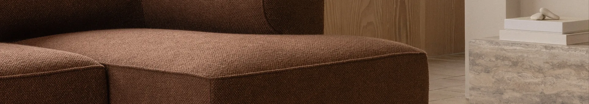 Close-up of a brown textured sofa, with a wooden wall and decorative objects on a concrete surface in the background.