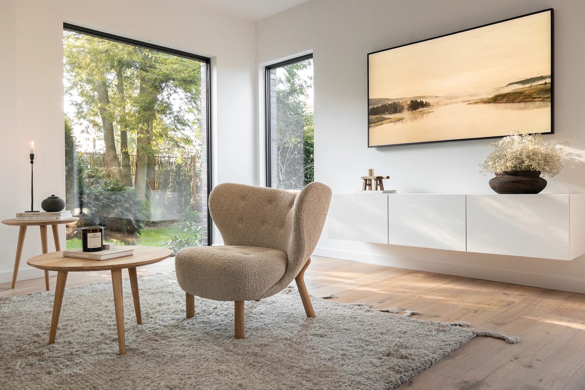 Modern living room with a cream bouclé armchair, two wooden coffee tables on a rug, a large window to a garden, and a TV showing a misty landscape.