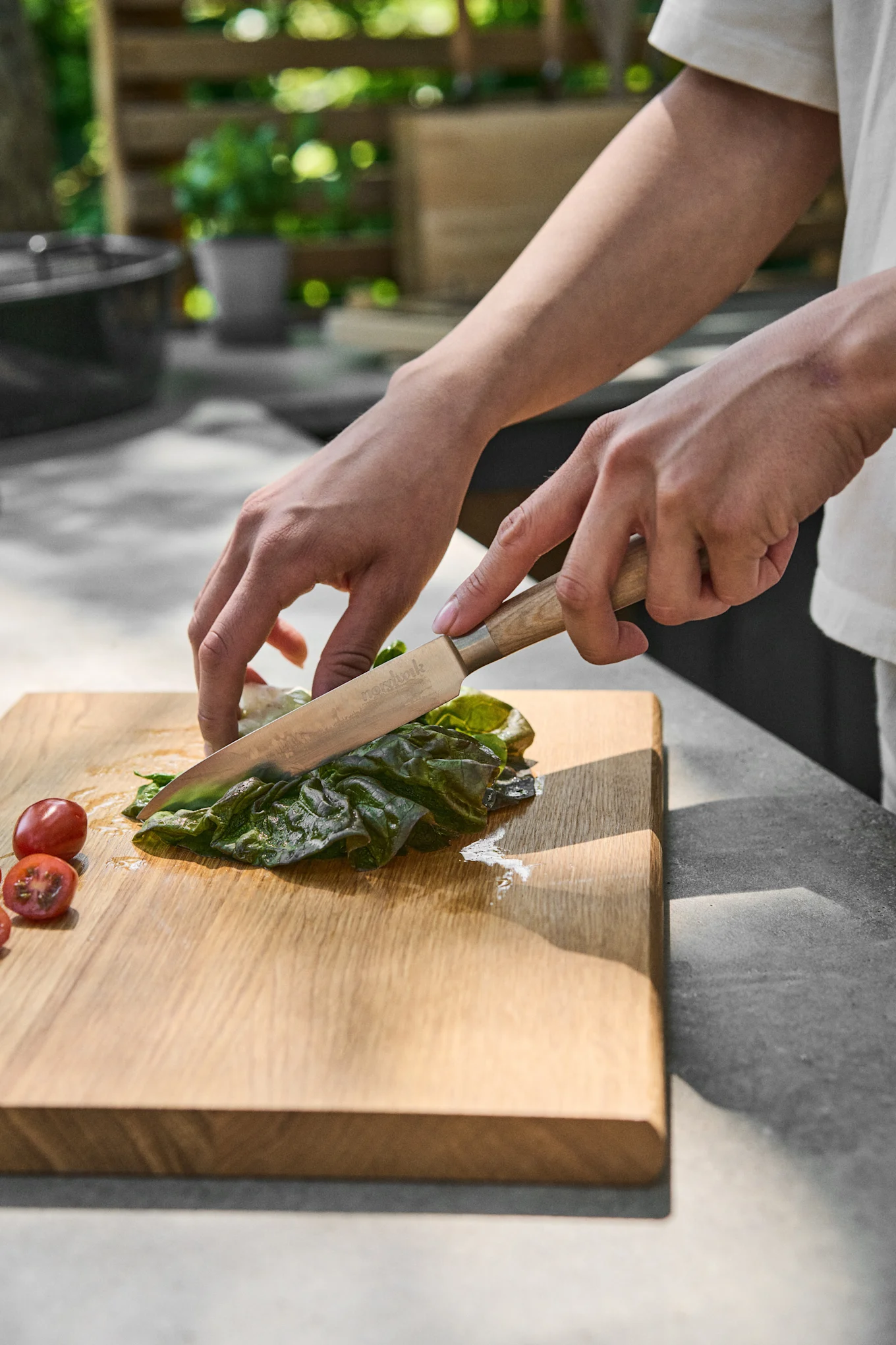 Hands slicing green leafy vegetables with a wooden-handled knife on a cutting board, with cherry tomatoes nearby.