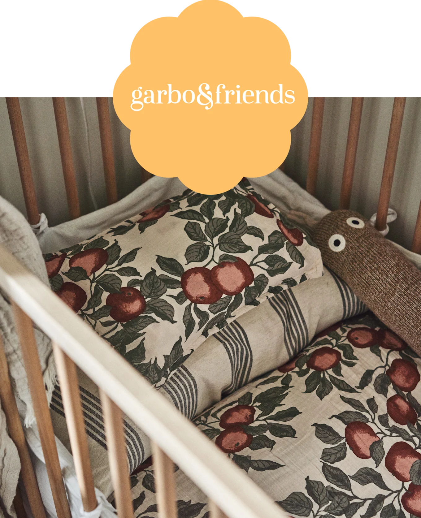 Crib bedding featuring red apple print and stripes, with a brown knitted toy. garbo&friends logo overlay.
