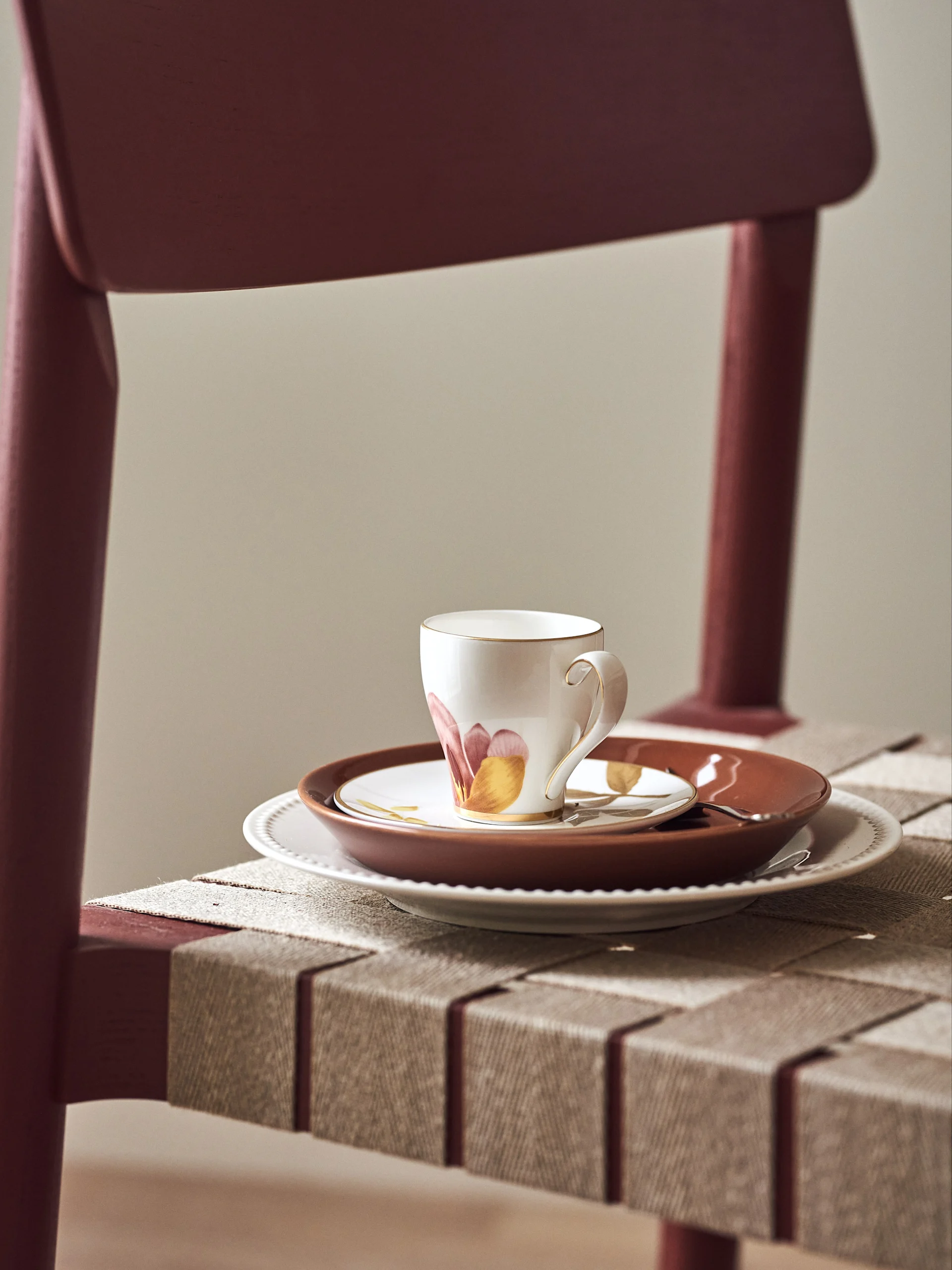 A white teacup with orange and red floral patterns and gold trim sits on stacked plates on a maroon woven chair.