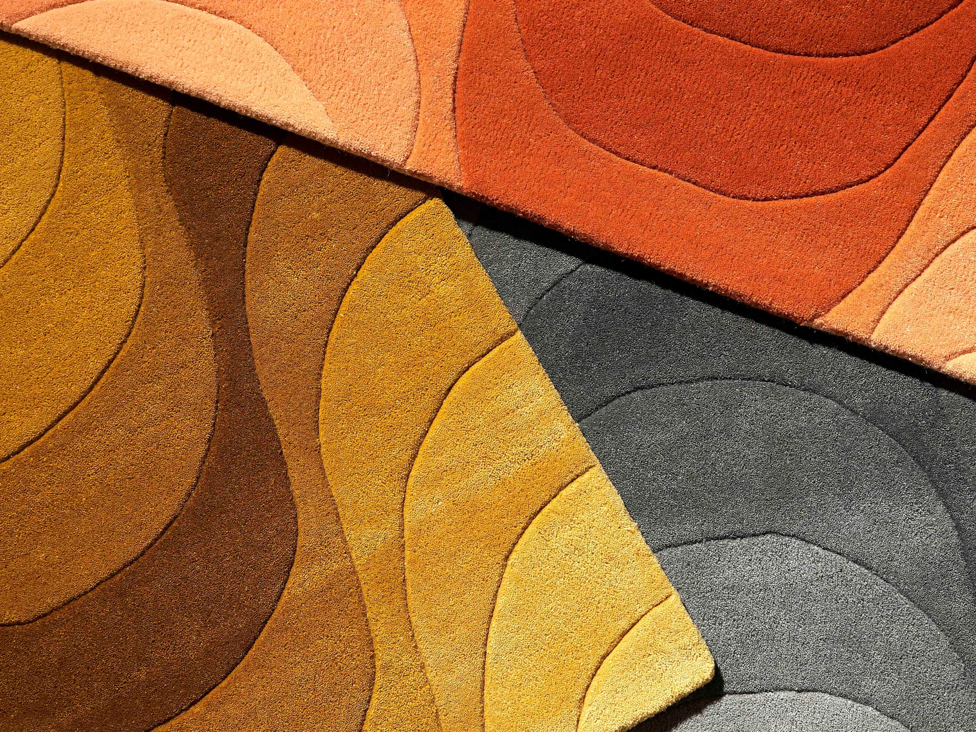 Close-up of several carpets with wavy patterns in shades of orange, yellow, brown, and dark grey.
