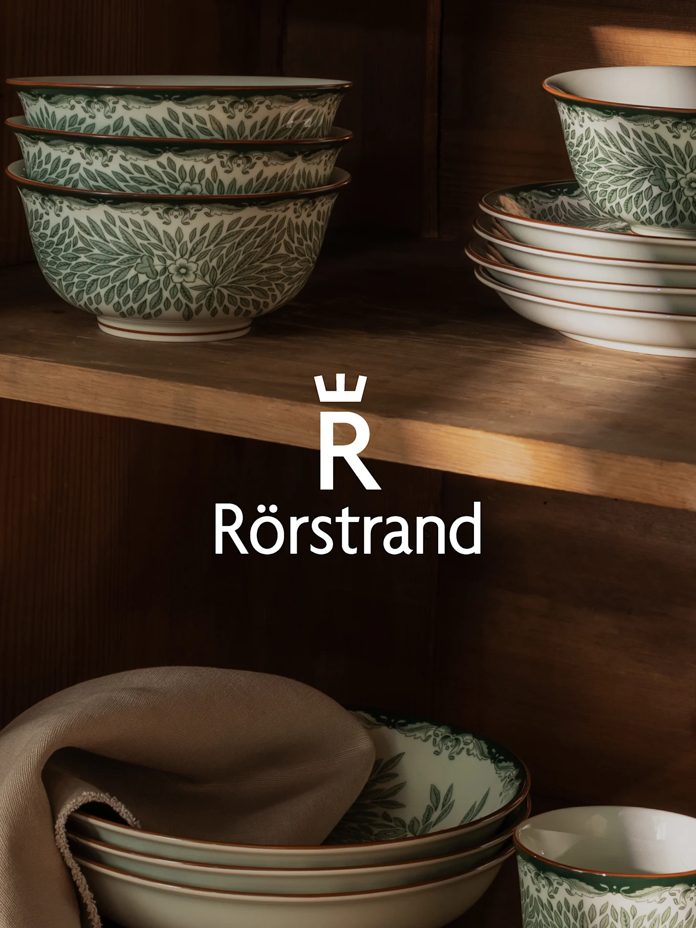 Stacked white bowls and plates with green leaf pattern on wooden shelves, featuring the Rörstrand logo.