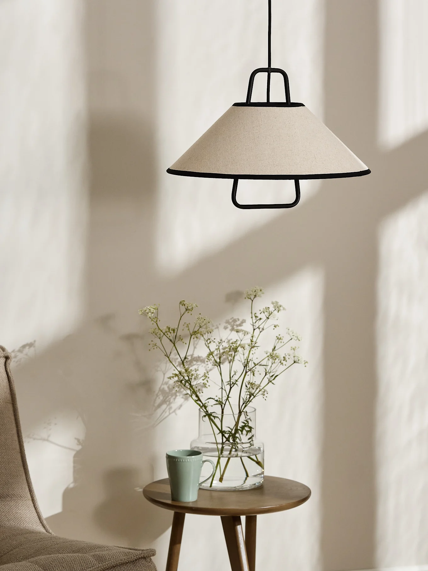 A conical cream pendant lamp with black trim hangs above a wooden side table with a vase of white flowers and a light green mug.