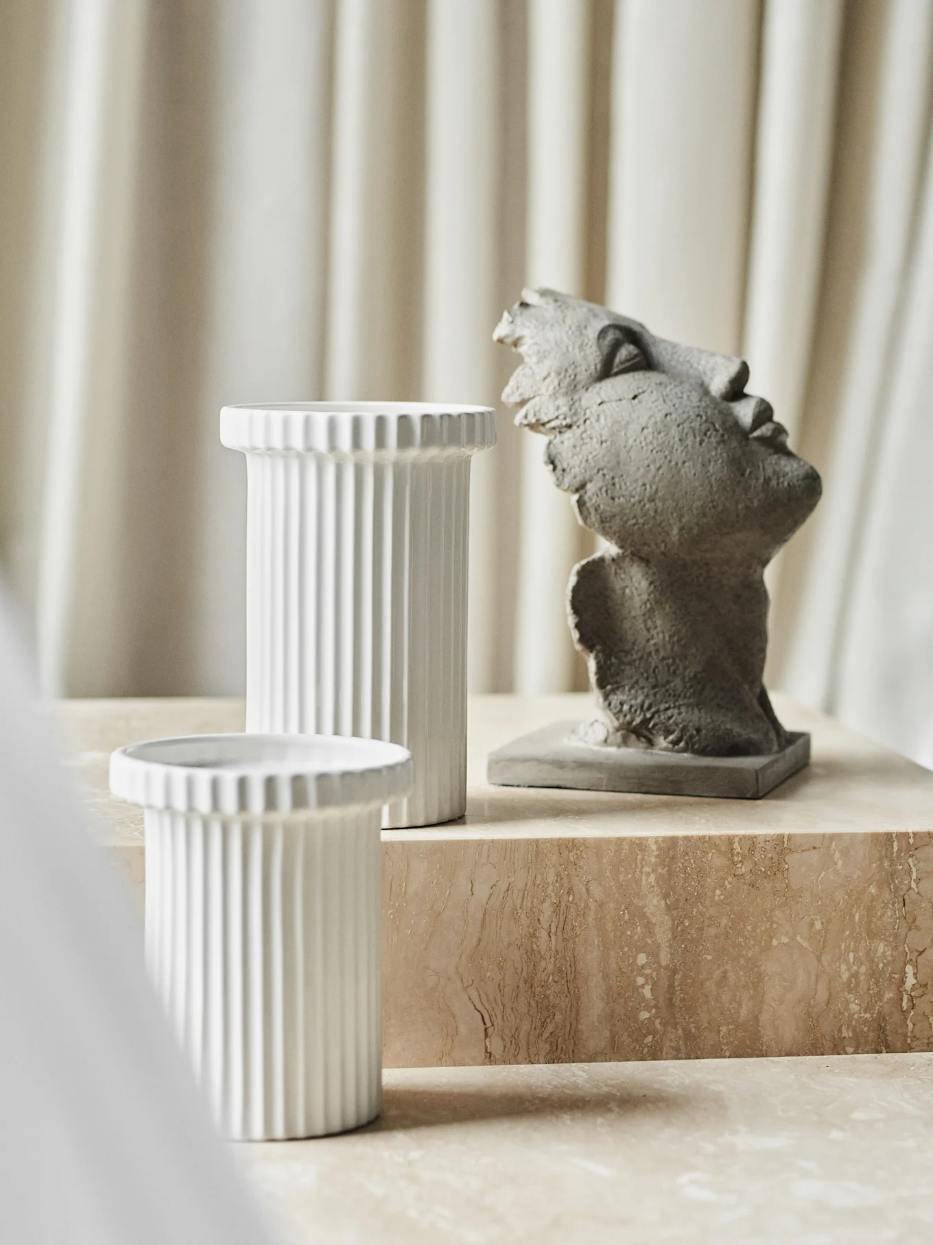 Two white ribbed cylindrical vases and a grey abstract head sculpture on a beige marble surface.