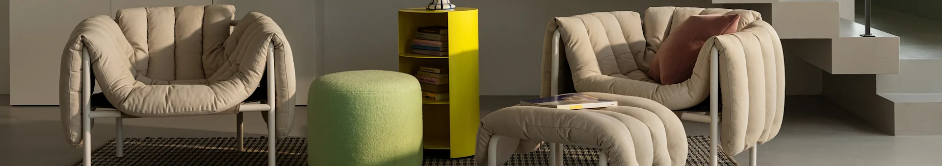 Two beige padded lounge chairs, a yellow shelf with books, and a green fuzzy ottoman in a modern room.