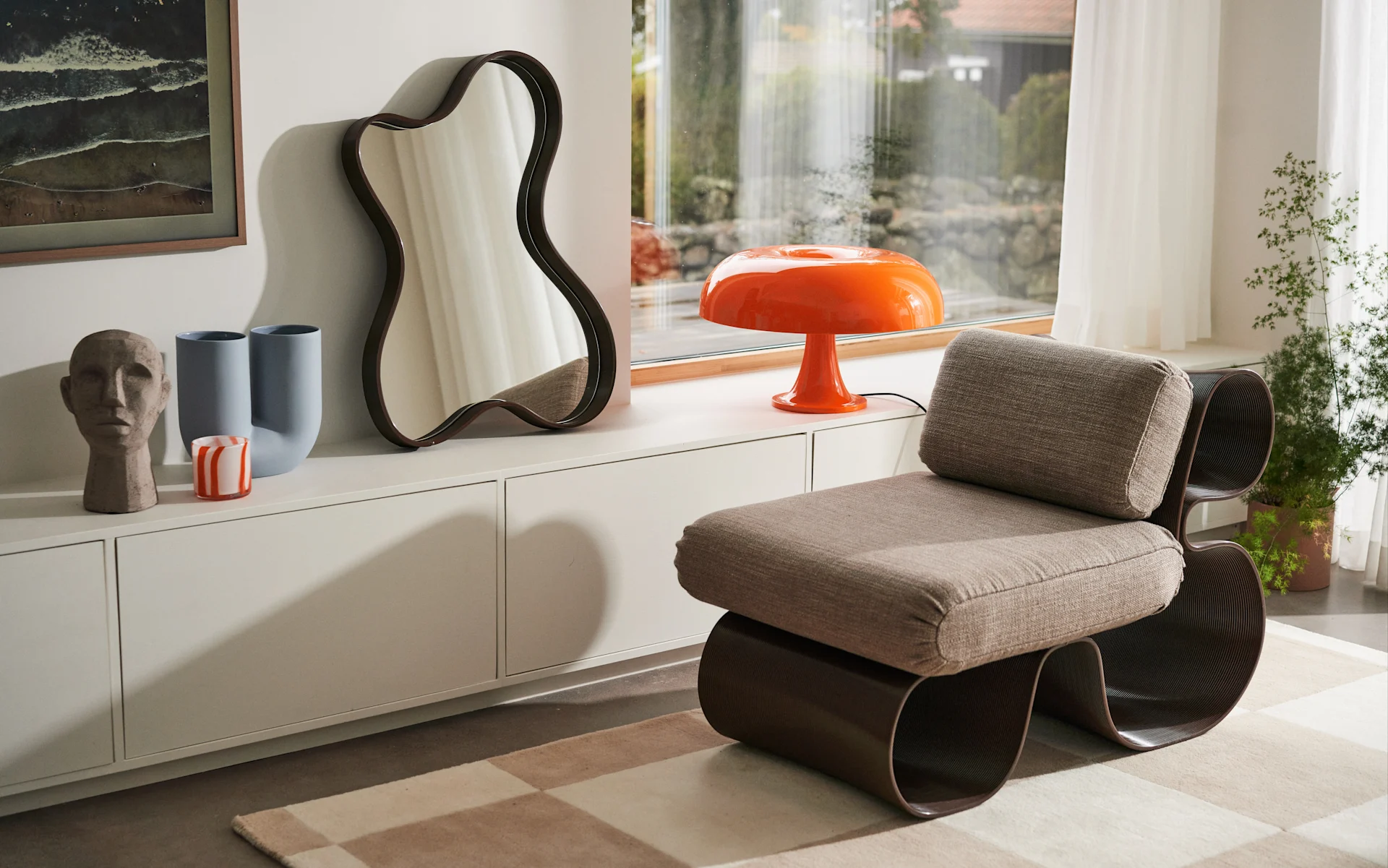 A modern room with a beige chair, orange mushroom lamp, wavy mirror, and vases on a white cabinet.