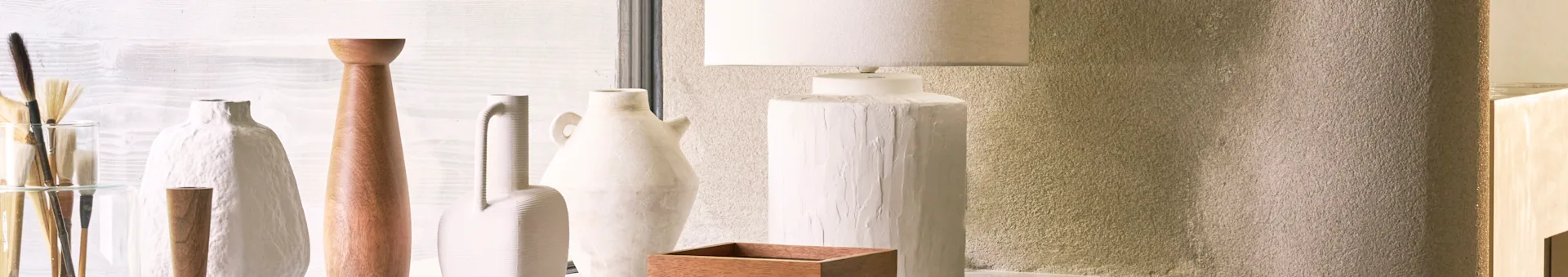 Decorative display with white and wooden vases, paintbrushes, and a textured white lamp on a shelf against a textured wall.