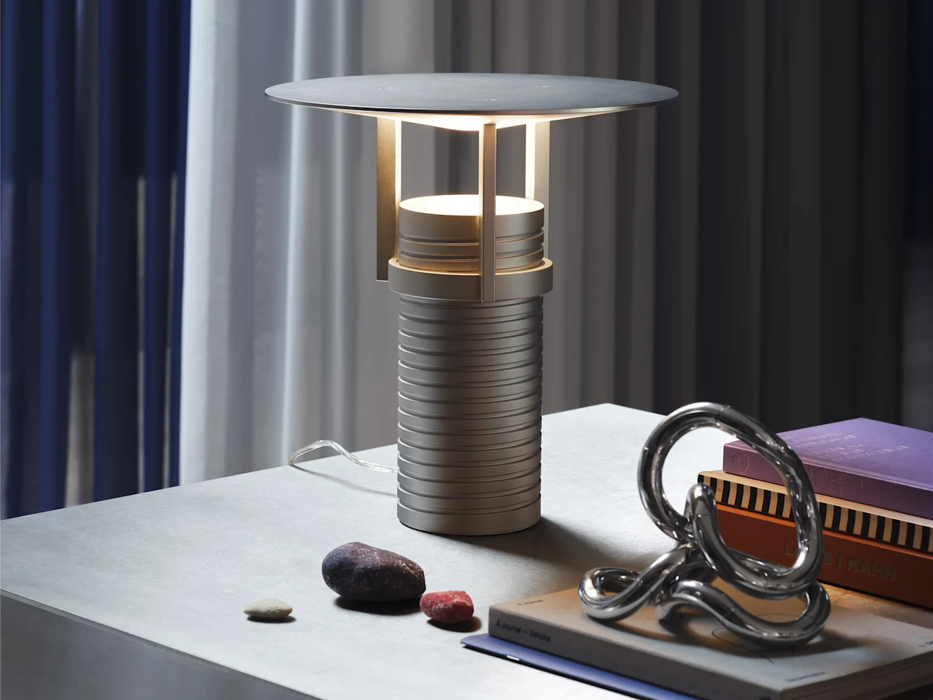 A modern grey table lamp with a ribbed base and circular shade, emitting a warm glow, sits on a grey table with decorative rocks, a silver sculpture, and books.