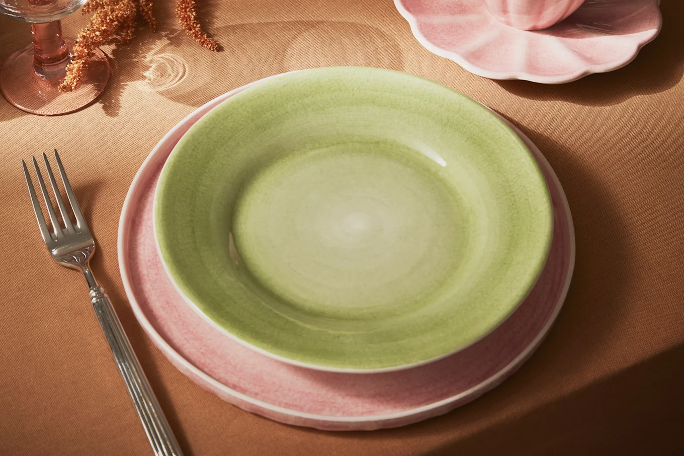 A green ceramic plate stacked on a pink one, with a silver fork, pink glass, and partial teacup on a brown tablecloth.