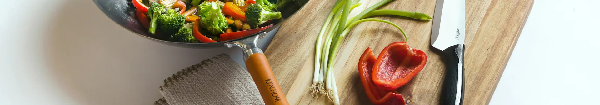 Wok of stir-fried vegetables (broccoli, peppers) next to a cutting board with spring onions and a knife.