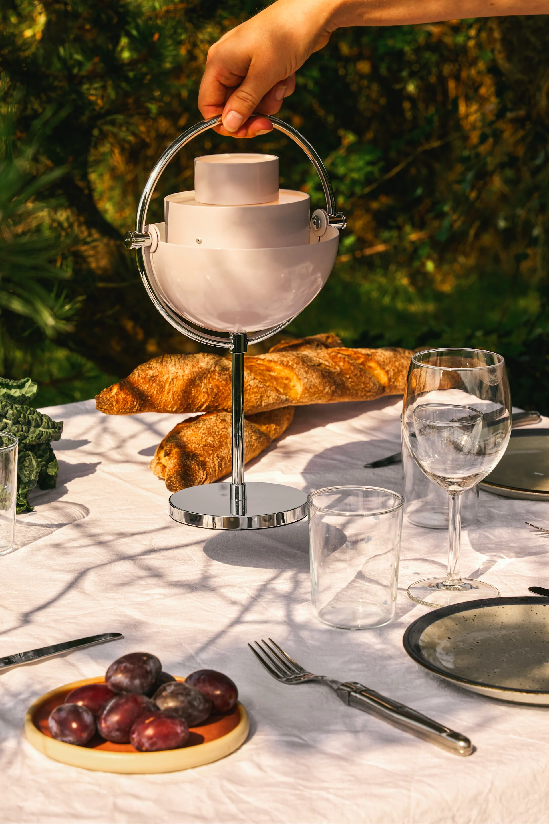Hand holds light pink, spherical, tiered serving dish with metal handle on an outdoor table set with food.