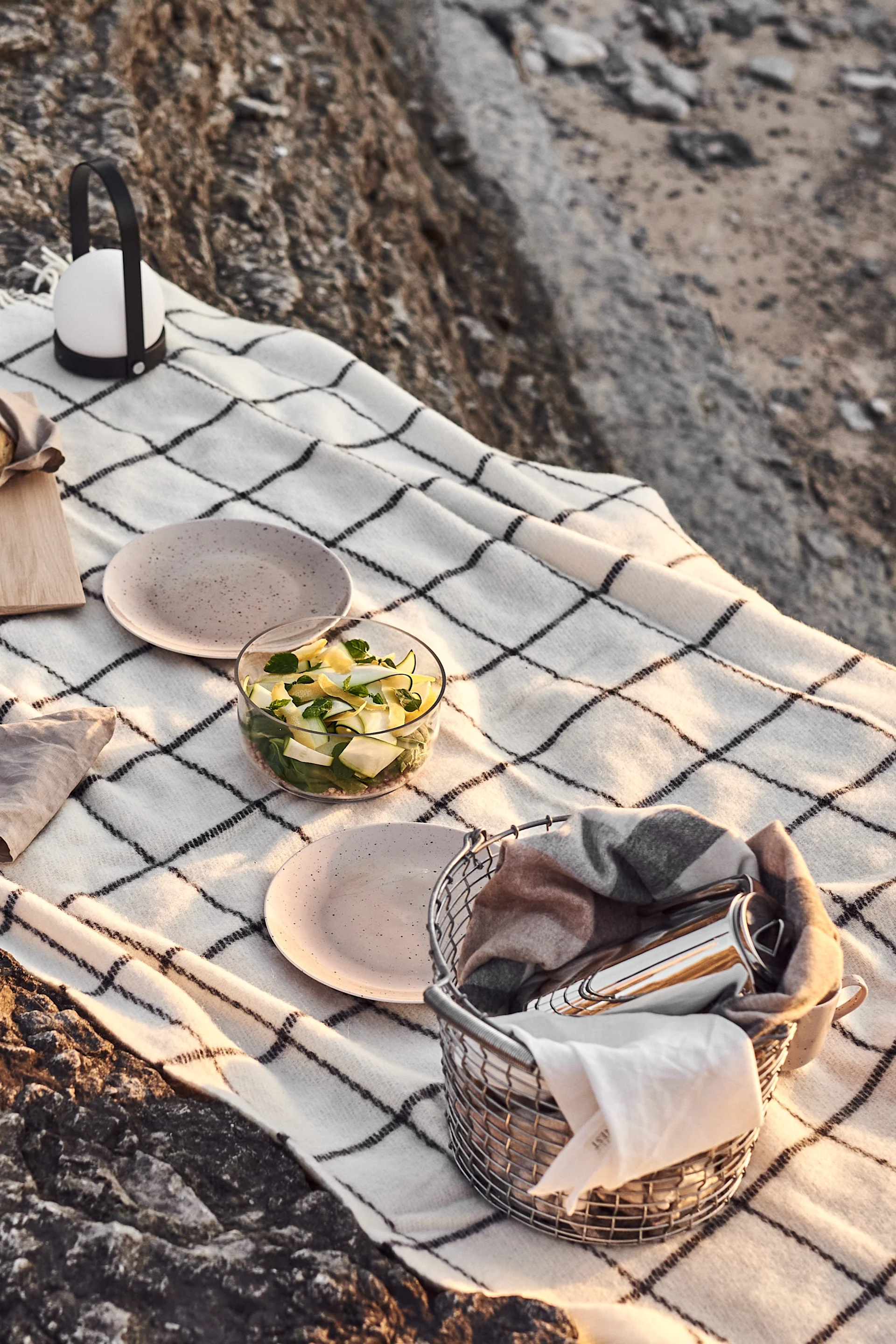 Picnic blanket on rocks with salad, plates, a lantern, and a basket of utensils.