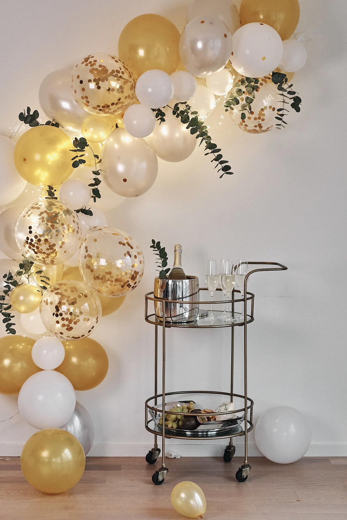Party scene with white and gold balloons, a gold bar cart with champagne and snacks, and string lights.