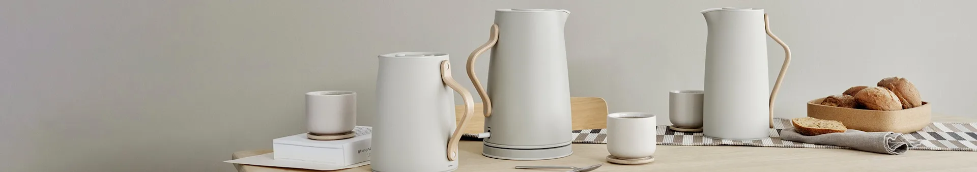 Minimalist table setting with three white kettles, matching mugs, and a bread basket on a wooden table.