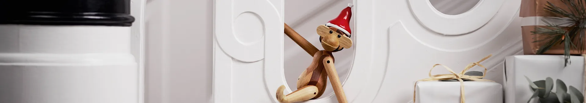 A wooden monkey toy with a red Santa hat peeking from behind white decor, with wrapped gifts in the background.