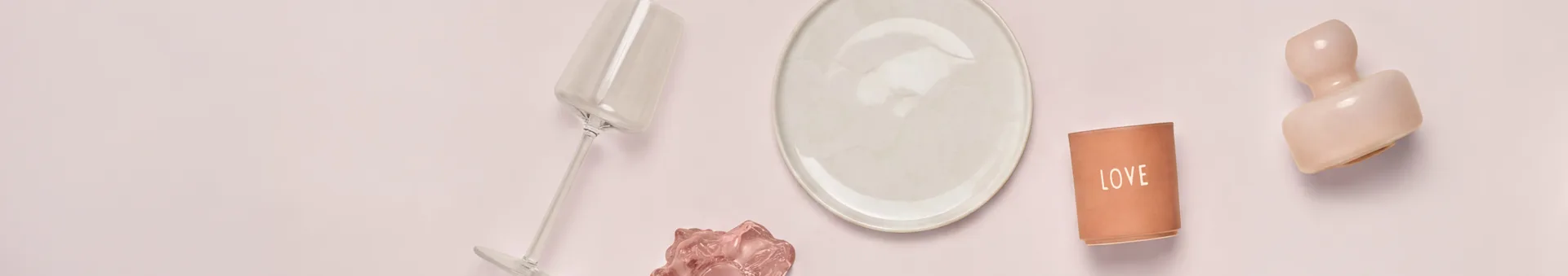A top-down view of various decorative and dining objects on a light pink surface. From left to right: a tilted clear glass, a cream plate, a terracotta "LOVE" candle holder, and a pink abstract sculpture. A smaller pink wavy object is below the glass.
