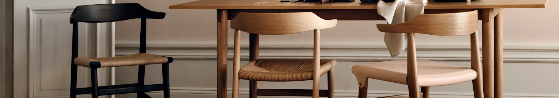 Three wooden chairs: a tall dark chair with a woven seat, a light wood chair with a woven seat, and a light wood chair with a pink upholstered seat, next to a table.