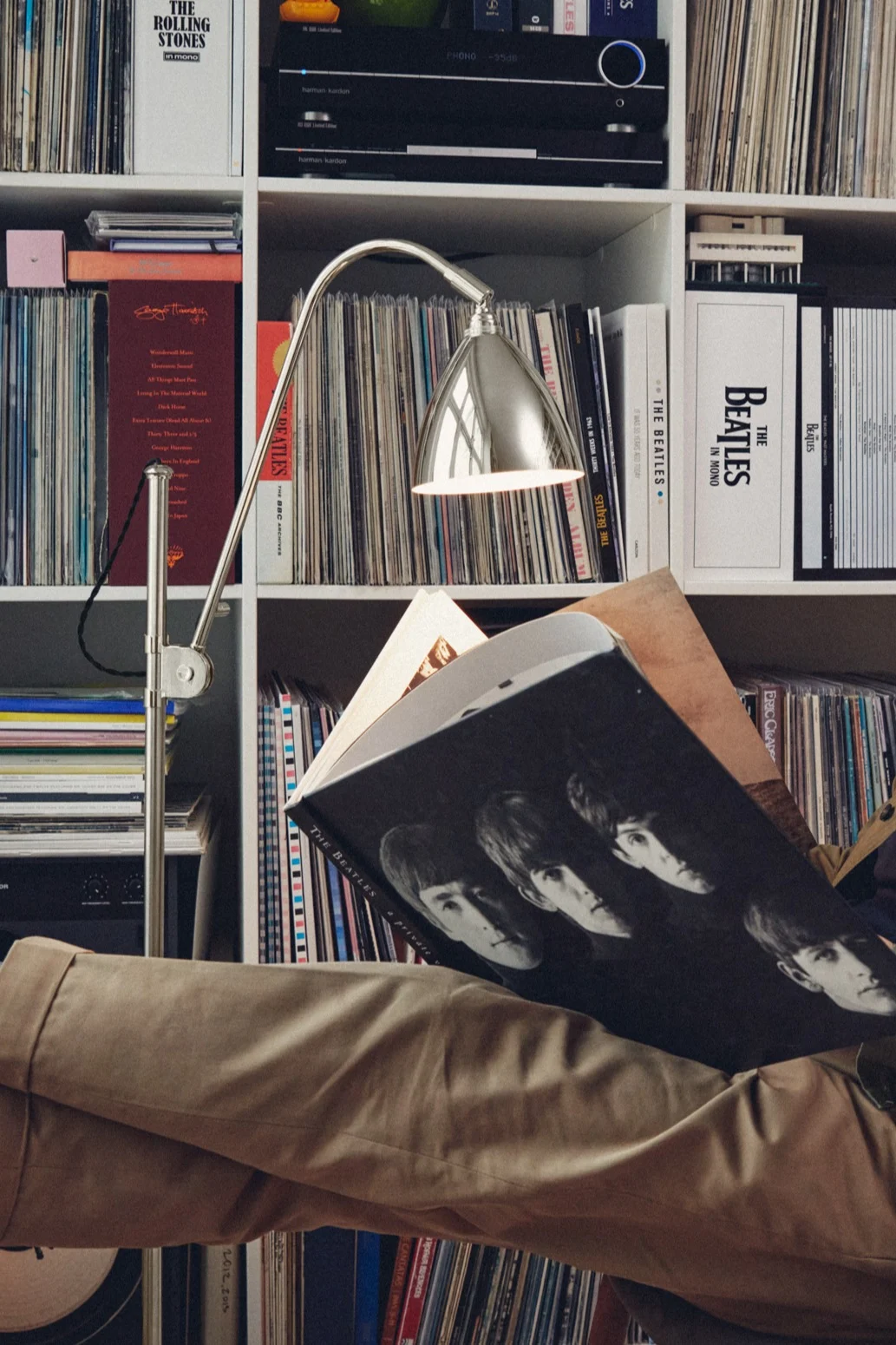 Person reading a book about The Beatles, illuminated by a silver lamp, in front of a shelf of vinyl records.