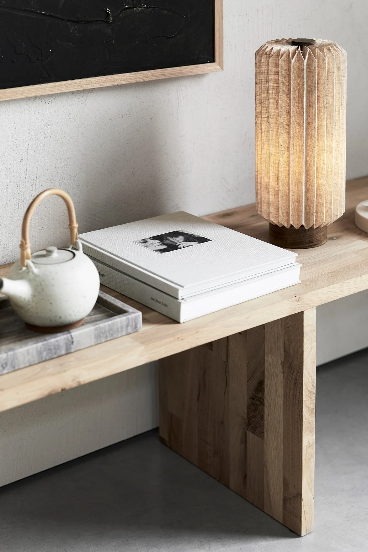 A wooden bench holds a teapot on a marble tray, stacked books with a photo, and a pleated fabric lamp.